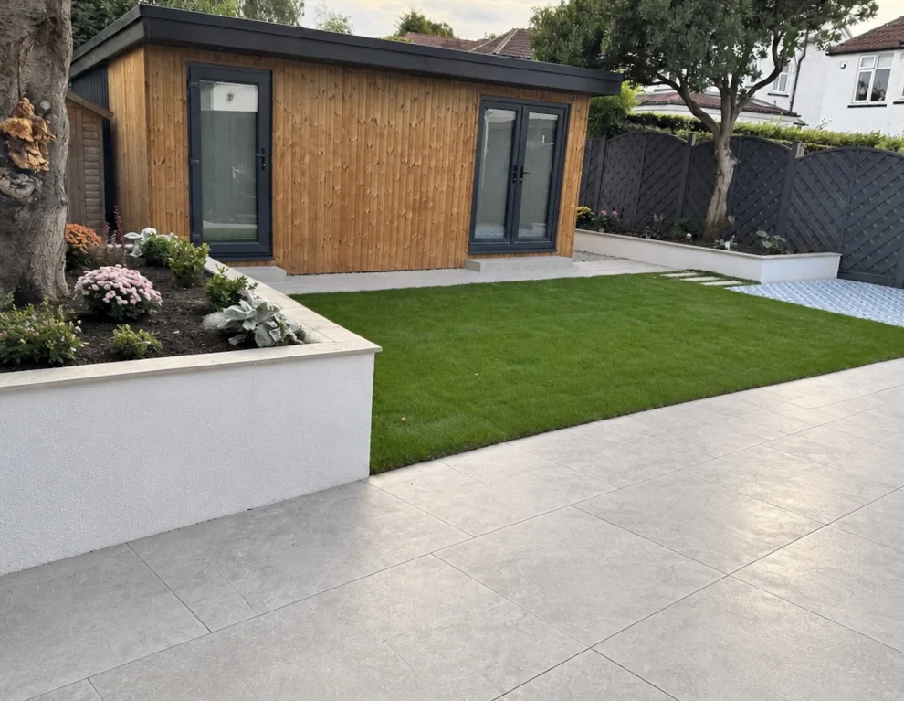 Light paving surrounding a rectangular lawn, with raised planted borders and a timber garden building